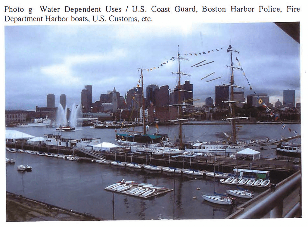 A panoramic view of a harbor with multiple boats docked, including sailing vessels, and a backdrop of a city skyline featuring several skyscrapers under a cloudy sky. Water fountains are visible in the harbor.