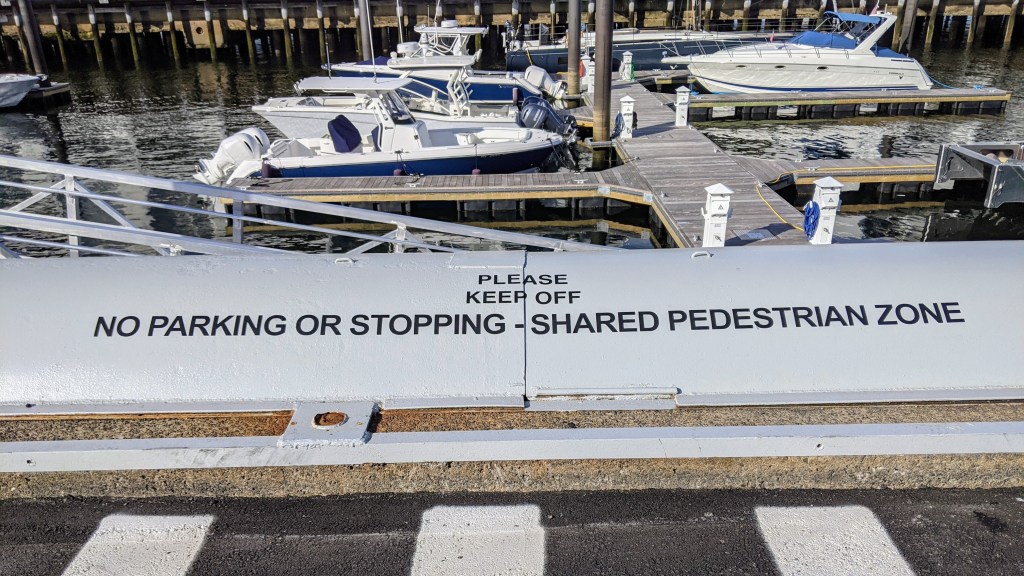 A sign at a marina stating 'PLEASE KEEP OFF NO PARKING OR STOPPING - SHARED PEDESTRIAN ZONE' with boats moored in the background.